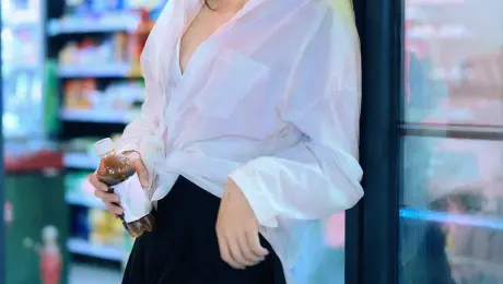 35mm film photography with harsh convenience store fluorescent lighting mixed with colorful neon signs from outside, authentic film grain, high contrast, slight color cast, cinematic street editorial style, intimate medium shot, early 20s sexy Chinese female idol with ultra-realistic delicate refined Chinese features, seductive almond-shaped fox eyes with natural double eyelids, high nose bridge, small sharp V-shaped jawline, flawless porcelain skin with cool ivory undertone and visible specular highlights from fluorescent light, subtle skin texture and micro pores, natural dewy makeup with soft flush on cheeks, glossy natural pink lips slightly parted, subtle natural freckles across nose and cheeks, long dark brown hair in a messy high ponytail with many loose strands falling around face and neck, wearing an oversized white button-up shirt as the only top, unbuttoned at the top with deep cleavage and loosely tied at the waist, paired with a tiny black pleated mini skirt, barefoot in simple white slides, seductive casual leaning pose against the glass door of a 24-hour convenience store at late night, body slightly arched, one leg bent with foot resting against the door frame, the other leg straight, one hand holding a bottle of iced drink, the other hand lightly pulling the hem of her mini skirt, intensely seductive playful yet slightly vulnerable gaze straight at the viewer with soft doe eyes full of quiet temptation and teasing smile, bright cold fluorescent store light from inside mixed with pink and blue neon glow from outside signs, realistic reflections on glass door, blurred convenience store interior with shelves and snacks in background, authentic 35mm film color grading with harsh lighting and neon accents, extremely sharp yet soft skin rendering, natural hair strands, realistic fabric wrinkles and drape on the oversized shirt and mini skirt, no plastic skin, no digital over-sharpening, no airbrushing, no blemishes, no moles, no oily skin, no watermark, no text, authentic late-night convenience store atmosphere