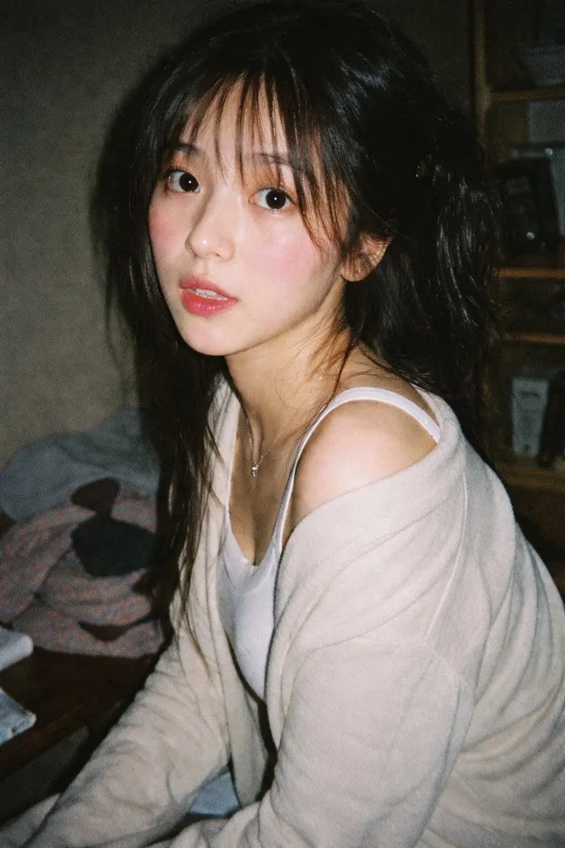 Generates a highly realistic, candid portrait with a vintage film aesthetic and direct flash lighting.
A vintage 35mm film photograph of a {argument name="subject description" default="young Asian woman"} with {argument name="hair style" default="long dark wavy hair and wispy bangs"}. She is wearing a {argument name="clothing" default="white ribbed tank top and a loose beige knit cardigan slipping off one shoulder"}, along with a delicate silver necklace. She has soft makeup with pink blush and glossy lips, looking directly at the camera with slightly parted lips. The lighting is harsh direct camera flash, creating a candid, amateur snapshot aesthetic. The background is a {argument name="setting" default="dimly lit, slightly messy room with clothes on a table and a wooden shelf"}. The image features heavy film grain, slightly muted colors, and a nostalgic, highly realistic photographic texture.
