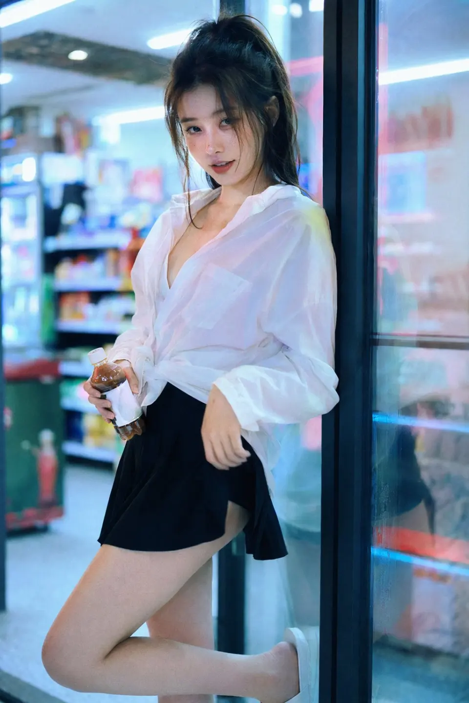 35mm film photography with harsh convenience store fluorescent lighting mixed with colorful neon signs from outside, authentic film grain, high contrast, slight color cast, cinematic street editorial style, intimate medium shot, early 20s sexy Chinese female idol with ultra-realistic delicate refined Chinese features, seductive almond-shaped fox eyes with natural double eyelids, high nose bridge, small sharp V-shaped jawline, flawless porcelain skin with cool ivory undertone and visible specular highlights from fluorescent light, subtle skin texture and micro pores, natural dewy makeup with soft flush on cheeks, glossy natural pink lips slightly parted, subtle natural freckles across nose and cheeks, long dark brown hair in a messy high ponytail with many loose strands falling around face and neck, wearing an oversized white button-up shirt as the only top, unbuttoned at the top with deep cleavage and loosely tied at the waist, paired with a tiny black pleated mini skirt, barefoot in simple white slides, seductive casual leaning pose against the glass door of a 24-hour convenience store at late night, body slightly arched, one leg bent with foot resting against the door frame, the other leg straight, one hand holding a bottle of iced drink, the other hand lightly pulling the hem of her mini skirt, intensely seductive playful yet slightly vulnerable gaze straight at the viewer with soft doe eyes full of quiet temptation and teasing smile, bright cold fluorescent store light from inside mixed with pink and blue neon glow from outside signs, realistic reflections on glass door, blurred convenience store interior with shelves and snacks in background, authentic 35mm film color grading with harsh lighting and neon accents, extremely sharp yet soft skin rendering, natural hair strands, realistic fabric wrinkles and drape on the oversized shirt and mini skirt, no plastic skin, no digital over-sharpening, no airbrushing, no blemishes, no moles, no oily skin, no watermark, no text, authentic late-night convenience store atmosphere