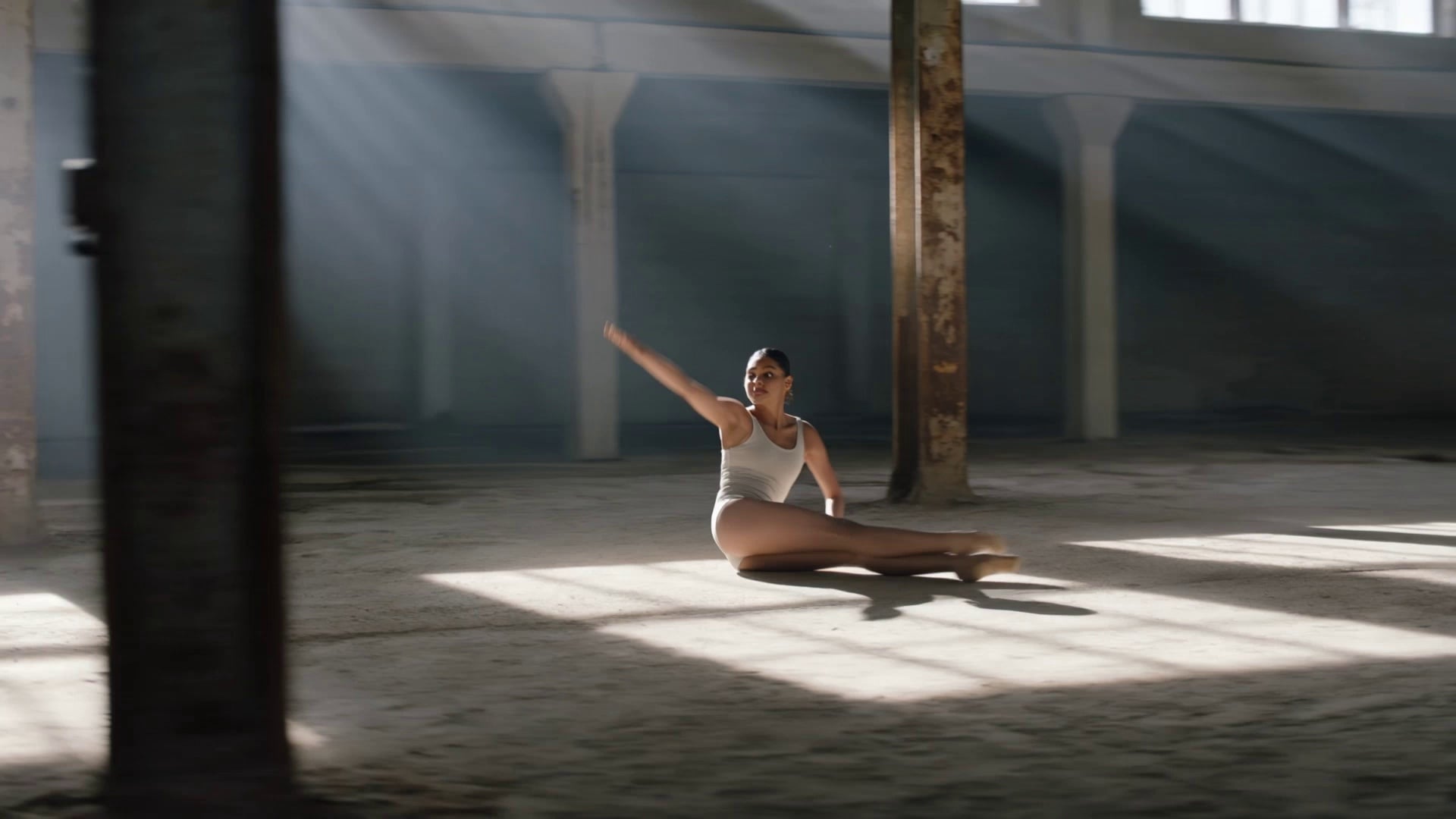 A contemporary dancer in minimalist attire performing a high-energy sequence of leaps and fluid floor work, kicking up subtle plumes of dust with every step inside a cavernous, abandoned concrete warehouse with rusted structural pillars. The camera executes a dynamic 360-degree orbital tracking shot that speeds up and slows down in synchronization with the choreography. Dramatic volumetric god rays pierce through high clerestory windows, creating high-contrast chiaroscuro shadows and illuminating floating particles in the air. Cinematic 35mm film aesthetic, shallow depth of field with soft bokeh on the distant background, high dynamic range.