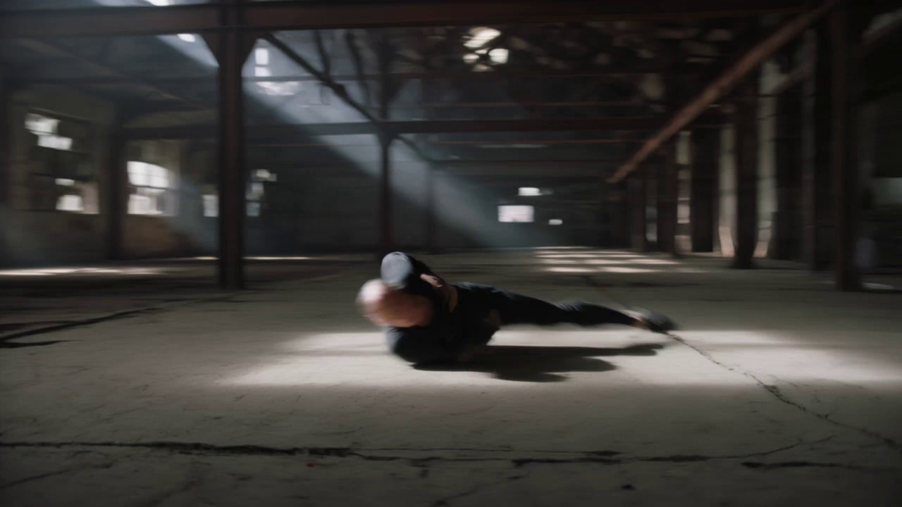 A contemporary dancer in fluid, monochromatic attire performing a high-energy sequence of leaps and floor work, kicking up plumes of dust from the cracked concrete. The scene takes place within a cavernous abandoned warehouse featuring rusted iron rafters and broken windows. A dynamic 360-degree orbital camera movement circles the performer at mid-range, capturing the kinetic energy of the movements. The atmosphere is defined by dramatic volumetric light beams piercing through the ceiling, creating high-contrast noir shadows and illuminating swirling atmospheric particles. Shot in 4K with a shallow depth of field, cinematic motion blur, and a gritty, desaturated color grade.