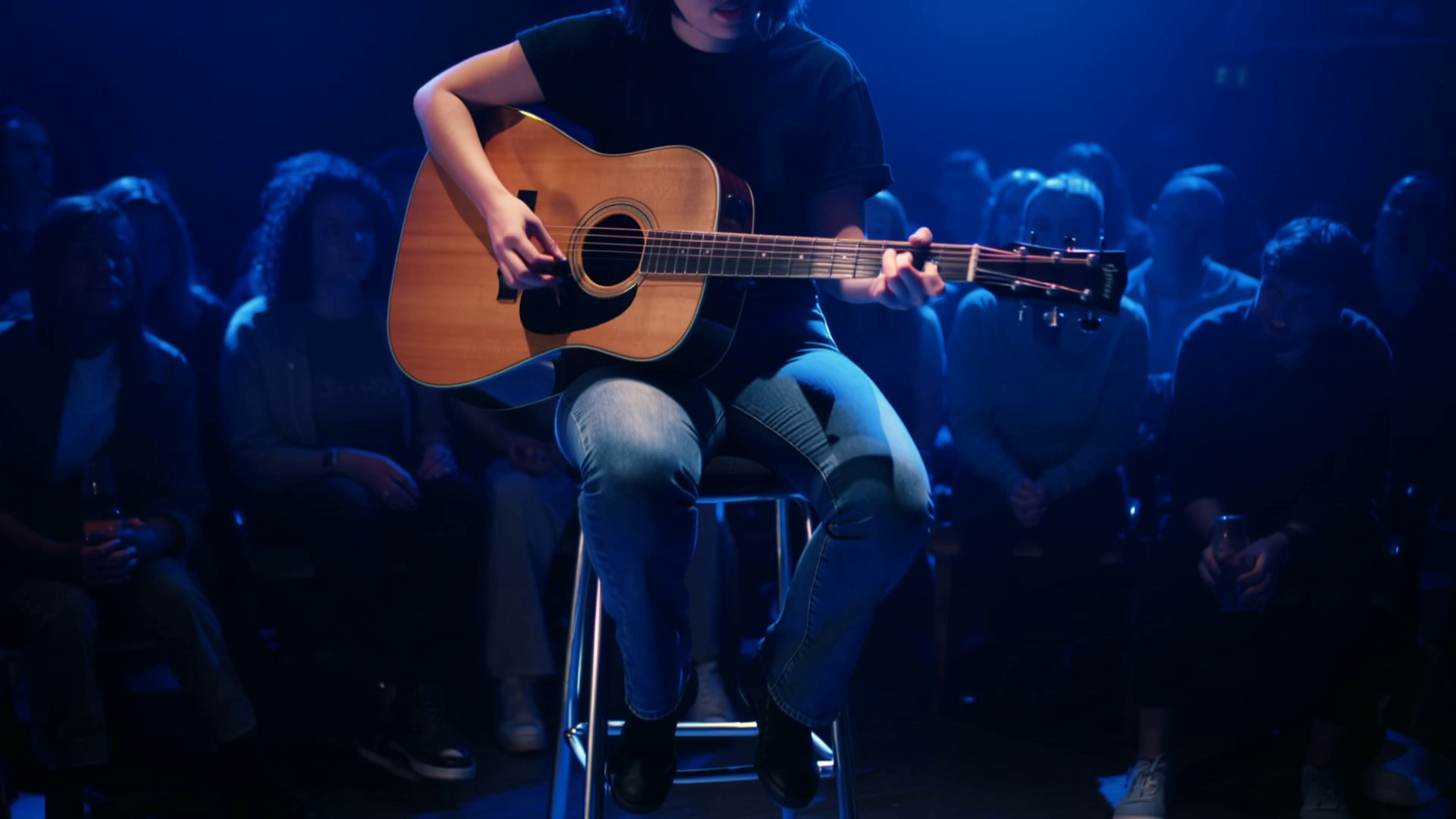 Scene: A livehouse venue with blue stage lights illuminating the performance area. A tall barstool is placed at the center of the stage, surrounded by an audience.
Subject: A short-haired female singer sits on the barstool, holding an acoustic guitar and performing live.
Audio: The short-haired female singer, emotional female voice, sings:
"And I will try to fix you, all night long..."
As she reaches the chorus, she looks out toward the audience.
Background includes soft audio feedback and the clinking of glasses.
Camera: Cuts between close-up shots of her fingers strumming the guitar and her expressive face as she sings.