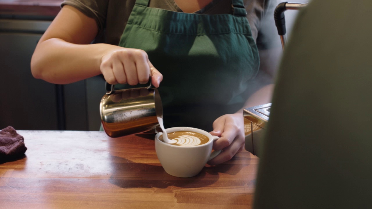 Candid style video of a busy barista working behind a counter in a crowded cafe. They are skillfully pouring latte art into a cup, then handing it to a customer with a quick smile. Steam rises from the machine. Natural movement and interactions.