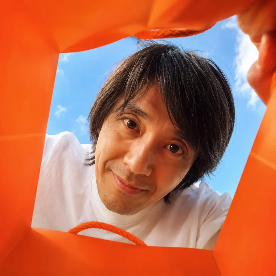 Create a vibrant, adorable, high-end advertising photo shot from inside a shopping bag, looking upward through the wide open top toward the sky.

The camera is placed deep inside the shopping bag, using a wide-angle lens at f/1.4 to create a playful close-up perspective with slight but charming wide-angle distortion. This distortion is important and must be clearly preserved, giving the image a fun, immersive, premium-advertising feel. The edges of the shopping bag should naturally frame the image, showing premium paper texture, soft folds, handles, and a clean commercial packaging aesthetic.

The shopping bag should be a high-saturation color, with the model randomly choosing a vivid tone such as hot pink, saturated yellow, blood orange, cobalt blue, vivid green, electric purple, deep cyan, or other bold commercial colors. The overall image should feel bright, cheerful, colorful, and visually striking.

Main subject:
If FACE_REF is uploaded:
Strictly preserve the uploaded person’s facial identity with 100% fidelity. Maintain the exact facial structure, eyes, eyelids, nose, lips, jawline, cheekbones, skin texture, age impression, hairstyle tendency, and overall identity. No beautification drift, no face reshaping, no age change, no ethnicity change, and no invented features.

If only PERSON_NAME is provided:
Create a highly recognizable portrait based on the public image of [周星驰], preserving their iconic facial features, hairstyle, expression tendency, posture, and personal aura as faithfully as possible.

The person leans deeply into the shopping bag from above, fully immersing their head into the opening as if curiously discovering something inside. Their face is very close to the camera lens, looking directly into it with wide, soulful, endearing eyes and a warm, adorable expression. The pose should feel playful, intimate, and heart-melting, as if they are curiously peeking into the bag and unexpectedly making eye contact with the viewer.

Lighting should feel bright, natural, and uplifting, with soft sunlight from the sky above. The image should maximize cuteness, charm, and “aww” factor while still feeling like a polished luxury advertising photo. The final result should instantly make the viewer smile.