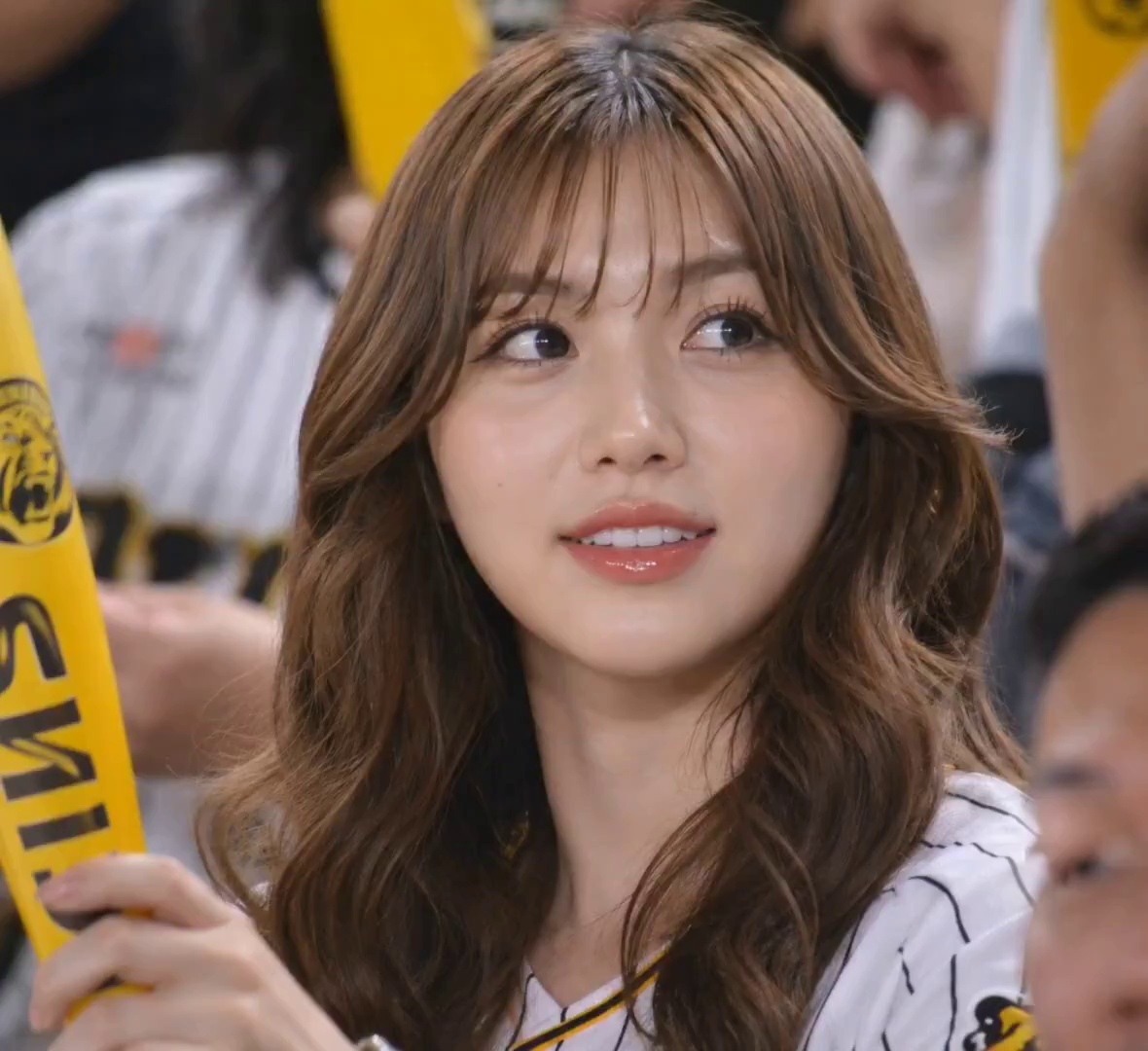 Use the uploaded reference image as the strongest identity anchor. The woman must look like the exact same adult Japanese woman from the reference image, not a similar person. Preserve her exact facial identity, same soft oval face, same glossy lips, same delicate nose, same large expressive eyes, same pale smooth skin, and same long voluminous curly blonde hair with soft layered bangs.

Create an ultra-realistic candid KBO baseball broadcast video scene during a lively night game. The woman is seated in the crowd beside her boyfriend, both wearing casual baseball jerseys among energetic cheering fans. She naturally watches the game while smiling and laughing softly with him. No dialogue, no talking, no cinematic acting. The interaction should feel completely natural and unscripted, like a real live broadcast moment accidentally captured by the stadium camera.

She holds yellow cheering sticks and lightly taps them together while cheering for the team. The boyfriend leans closer and they share a very short soft kiss lasting about one second, subtle and natural, not dramatic or romanticized. Immediately after the kiss, she realizes the live stadium camera is focused on them on the giant screen.

She becomes shy and slightly embarrassed for a moment, then gives a soft cute smile toward the camera while trying not to laugh. Her reaction should feel authentic, candid, and spontaneous, like a real fan unexpectedly shown on TV.

Use realistic Korean baseball TV broadcast cinematography: long telephoto lens compression, slight handheld camera movement, subtle motion blur from cheering fans, realistic stadium lighting, shallow depth of field, natural skin texture, broadcast softness, authentic crowd reactions, imperfect framing, 16:9 live sports broadcast composition, genuine candid atmosphere.