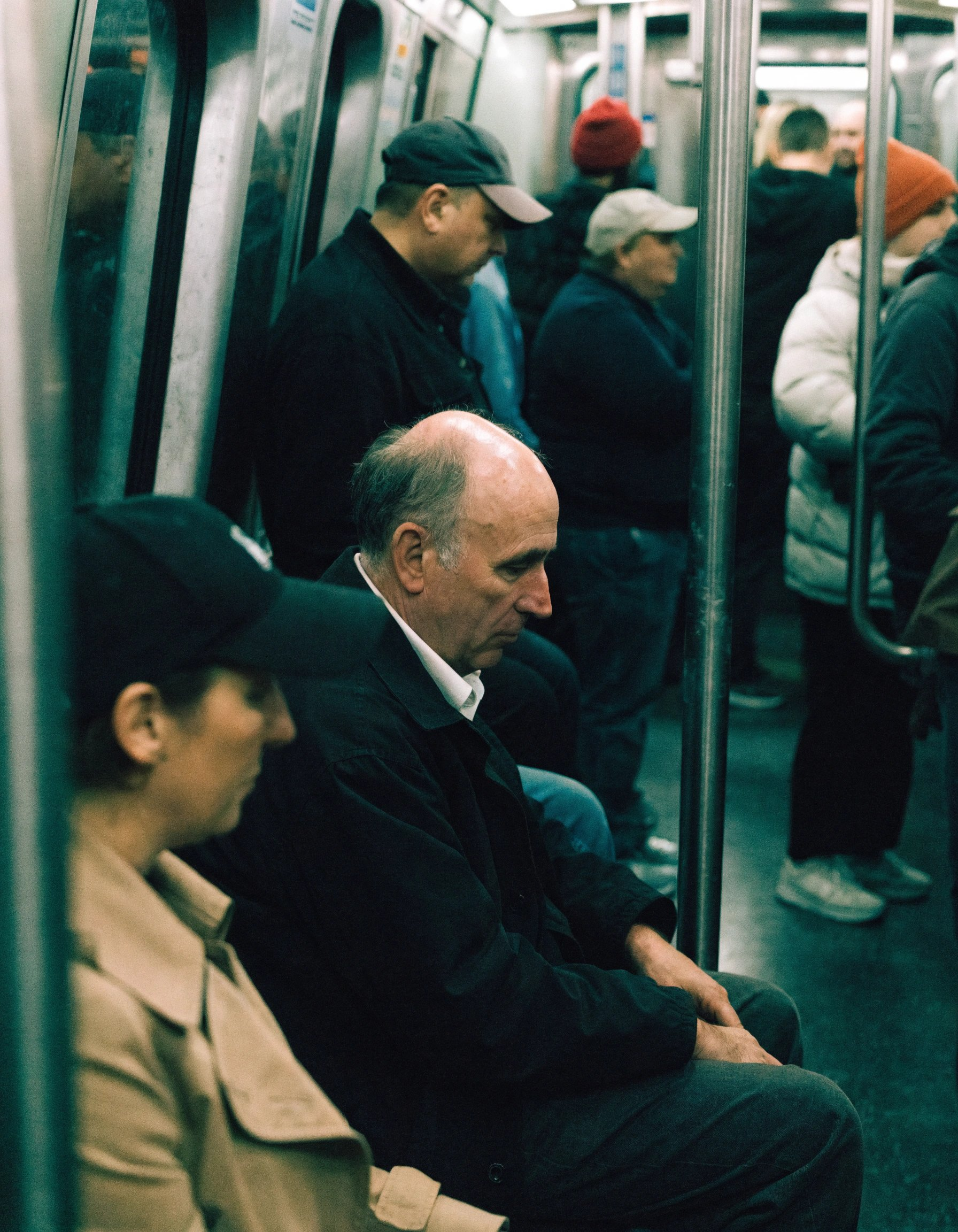 Interior of a subway train during a regular commute, a middle-aged man sitting among other passengers, wearing a simple jacket and shirt, leaning slightly and looking tired or lost in thought, completely natural posture with no awareness of the camera. Everyday, unstyled clothing with slightly worn textures. Harsh mixed subway lighting with greenish fluorescent tones, uneven illumination, and subtle color shifts. Candid handheld shot from a nearby seat, partially obstructed by poles and other passengers, with imperfect framing and slightly soft focus. The environment is crowded and tight, with overlapping bodies, blurred faces, metal poles, plastic seats, reflections, and worn surfaces. The scene feels intimate and observational, capturing quiet fatigue and routine — a relatable, unpolished moment of daily life.