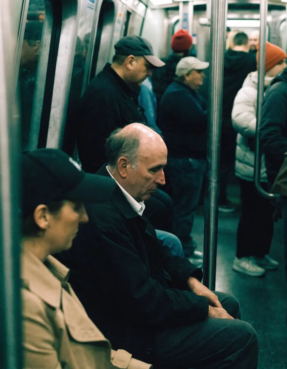 Interior of a subway train during a regular commute, a middle-aged man sitting among other passengers, wearing a simple jacket and shirt, leaning slightly and looking tired or lost in thought, completely natural posture with no awareness of the camera. Everyday, unstyled clothing with slightly worn textures. Harsh mixed subway lighting with greenish fluorescent tones, uneven illumination, and subtle color shifts. Candid handheld shot from a nearby seat, partially obstructed by poles and other passengers, with imperfect framing and slightly soft focus. The environment is crowded and tight, with overlapping bodies, blurred faces, metal poles, plastic seats, reflections, and worn surfaces. The scene feels intimate and observational, capturing quiet fatigue and routine — a relatable, unpolished moment of daily life.
